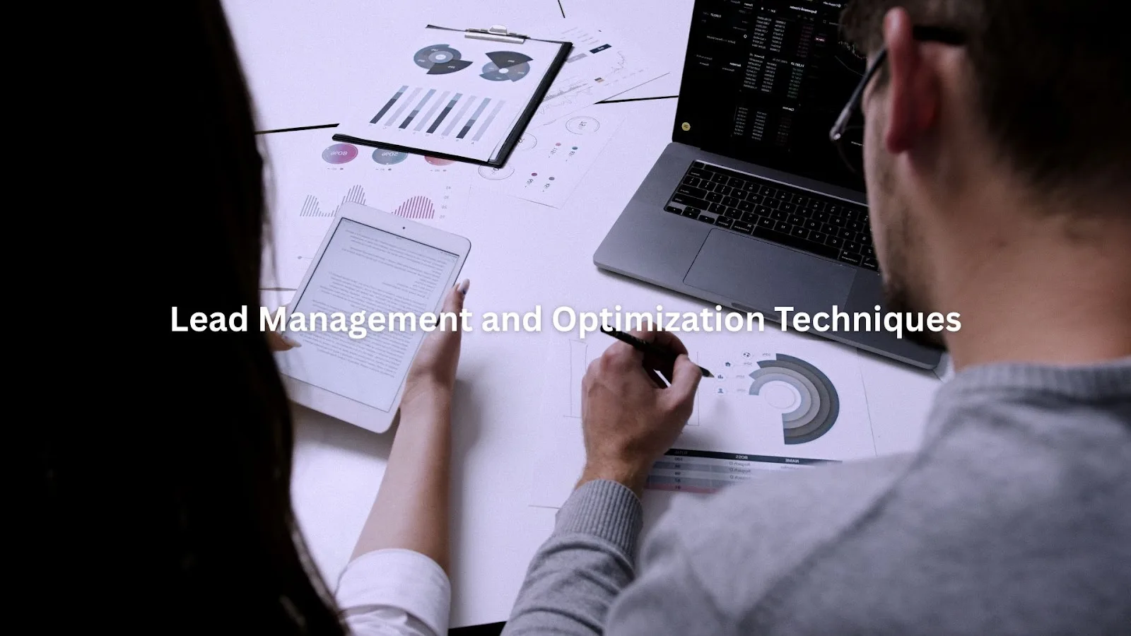 Two professionals analyzing charts and data on a desk with a laptop and tablet during a lead management meeting.